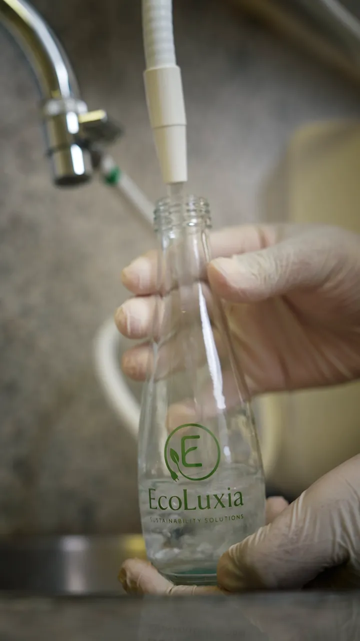 An EcoLuxia glass bottle filled under the K8’s wand, the glass gallon set on a dispenser, a glass poured and a bottle capped.