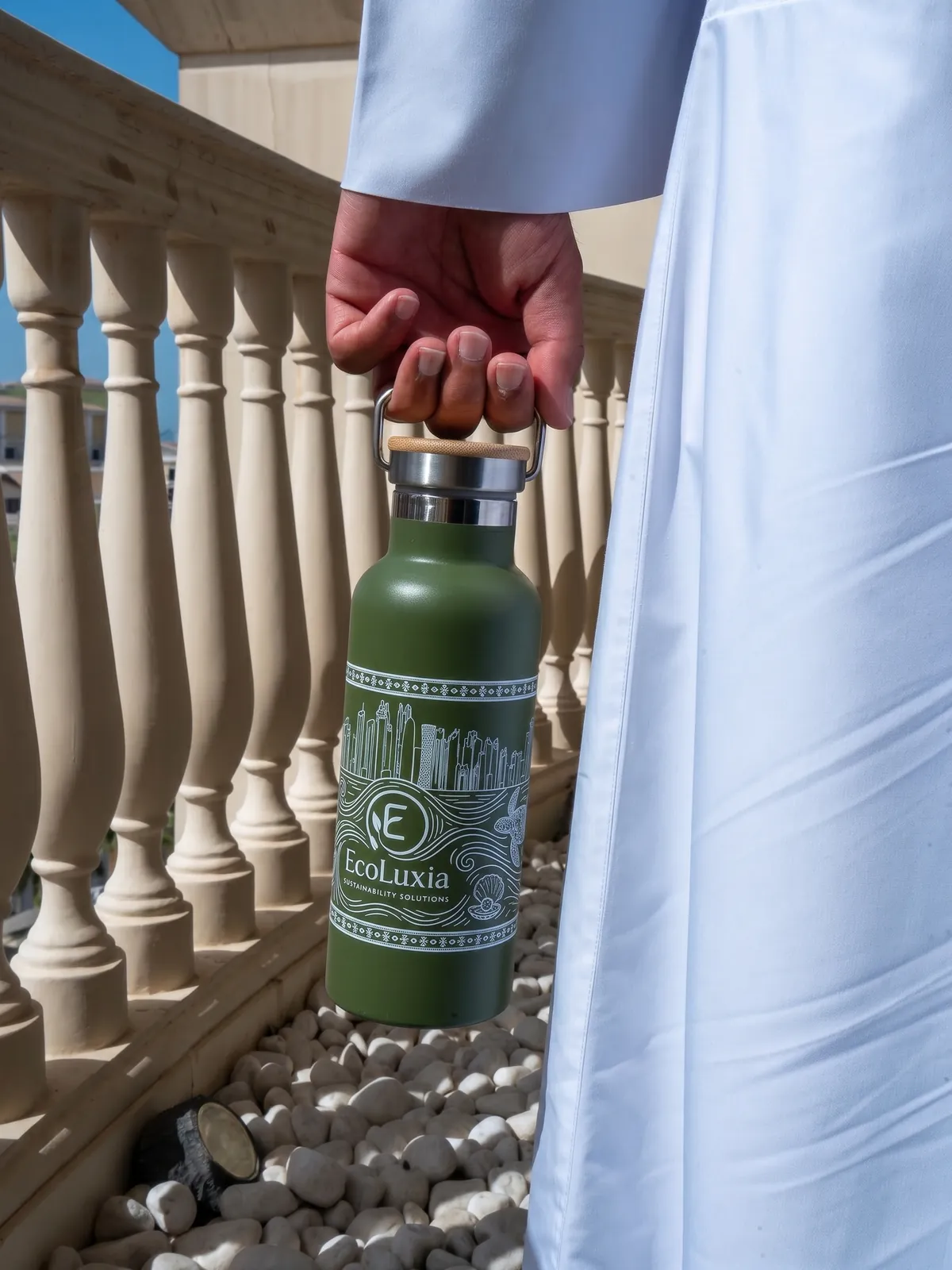 An EcoLuxia stainless-steel bottle carried by its handle beside a stone balustrade.