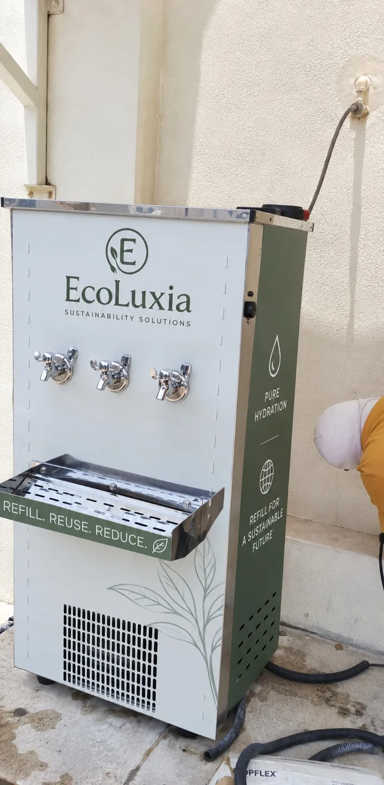 The freestanding EcoLuxia refill unit, with three taps over a drip tray and the words “Refill. Reuse. Reduce.”, installed outdoors against a wall.