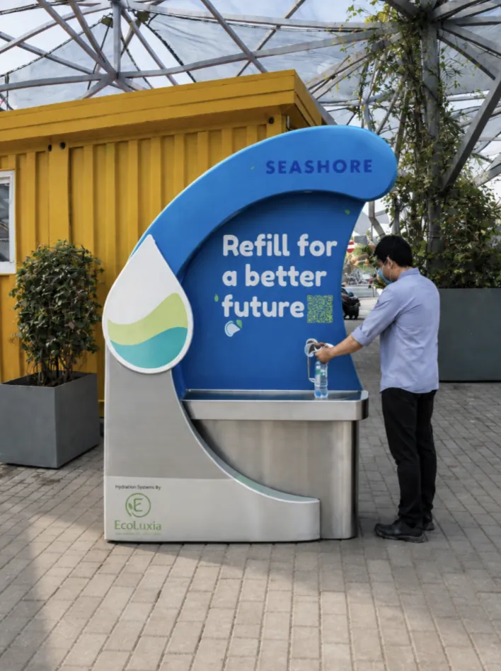 The Seashore refill unit at Green Island, Qatar Foundation: a curved blue panel reading “Refill for a better future”, with EcoLuxia’s logo at its base, as a visitor fills a bottle.